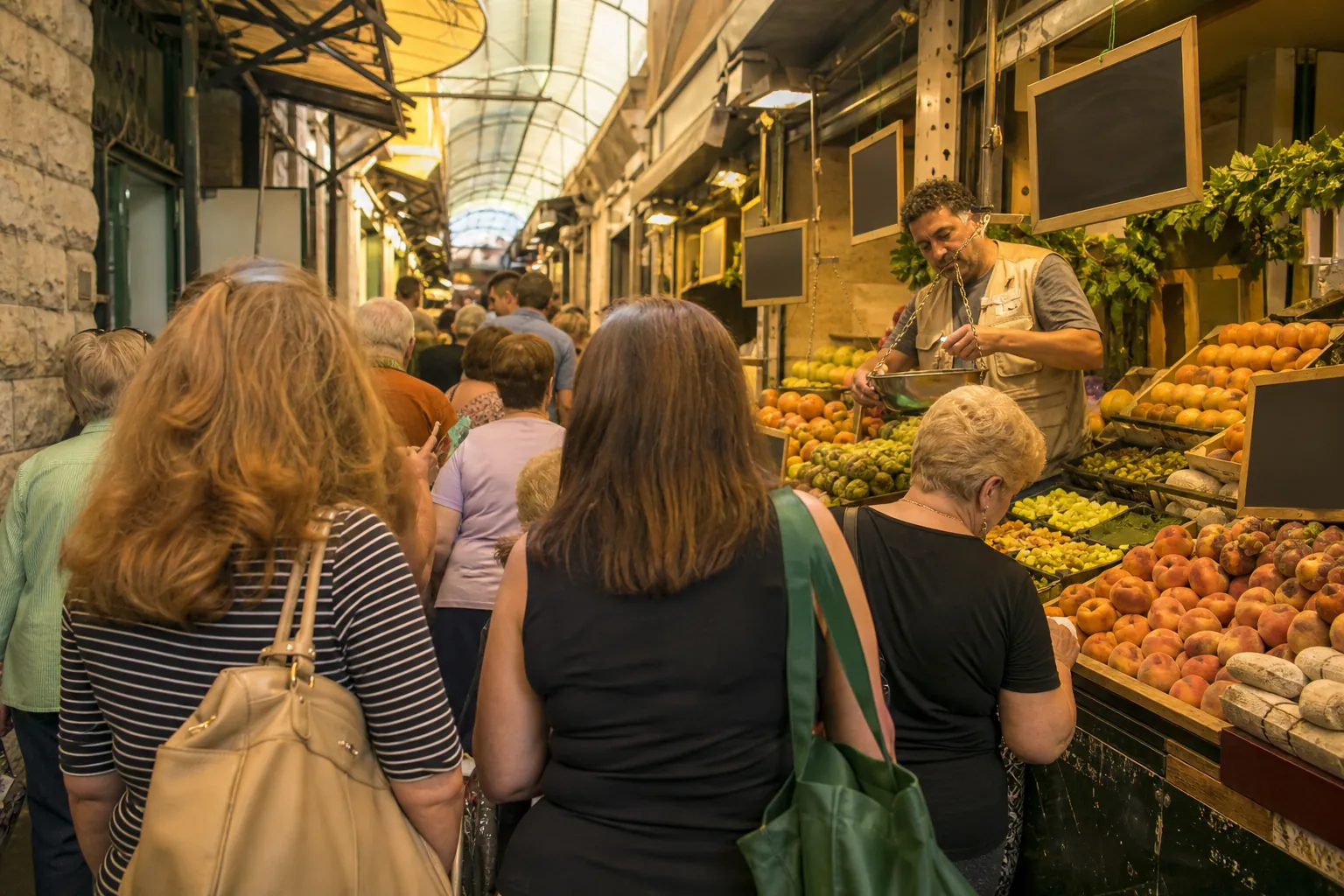 View from the back of a short queue toward a busy produce stall, shoppers studying piles of fruit and handwritten signs, seller weighing goods on a hanging scale, shoulder bags brushing against narrow aisle walls, 1200x800